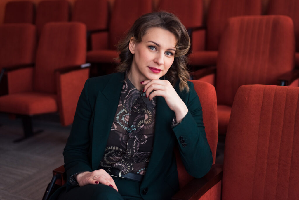 Portrait Of Beautiful Young Woman Sitting At Velvet Seats In Conference Hall Or Theatre, Looking At Camera