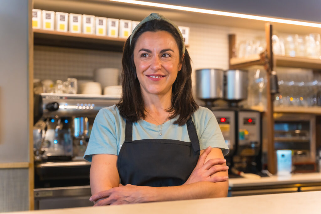 Portrait Of A Smiling Waitress With The Coffee Machine In The Background, The Covid Restrictions
