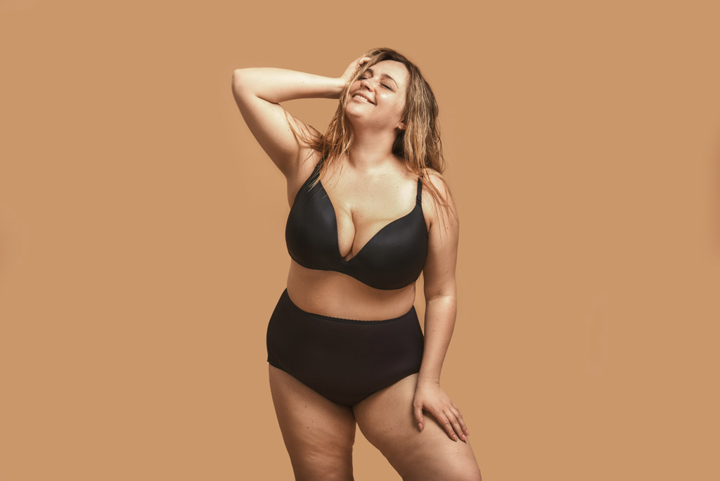Beautiful And Happy Plus Size Woman In Black Underwear Keeping Eyes Closed And Smiling While Standing In Studio Against Brown Background