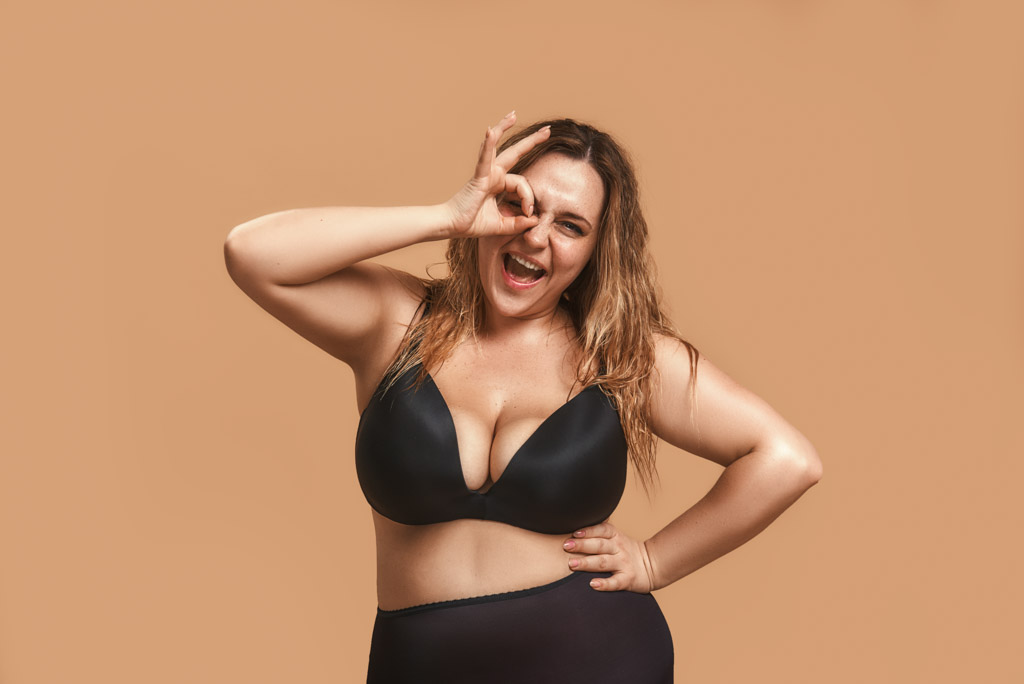 Everything Is Okay Happy Plump Woman In Black Underwear Showing Ok Sign With Fingers And Winking While Standing Against Brown Wall In Studio