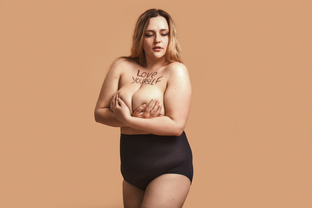 I Love My Body. Portrait Of Beautiful Plus Size Naked Woman Holding Her Bust With Written Text Love Yourself On It While Standing Against Brown Wall In Studio
