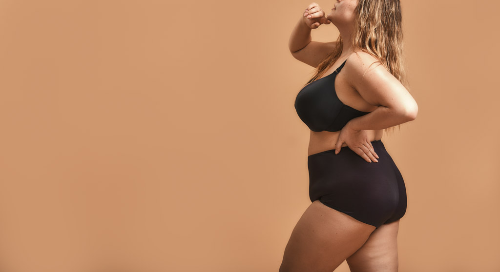 Side View Of Plus Size Sexy Model In Black Lingerie Standing Against Brown Wall In Studio