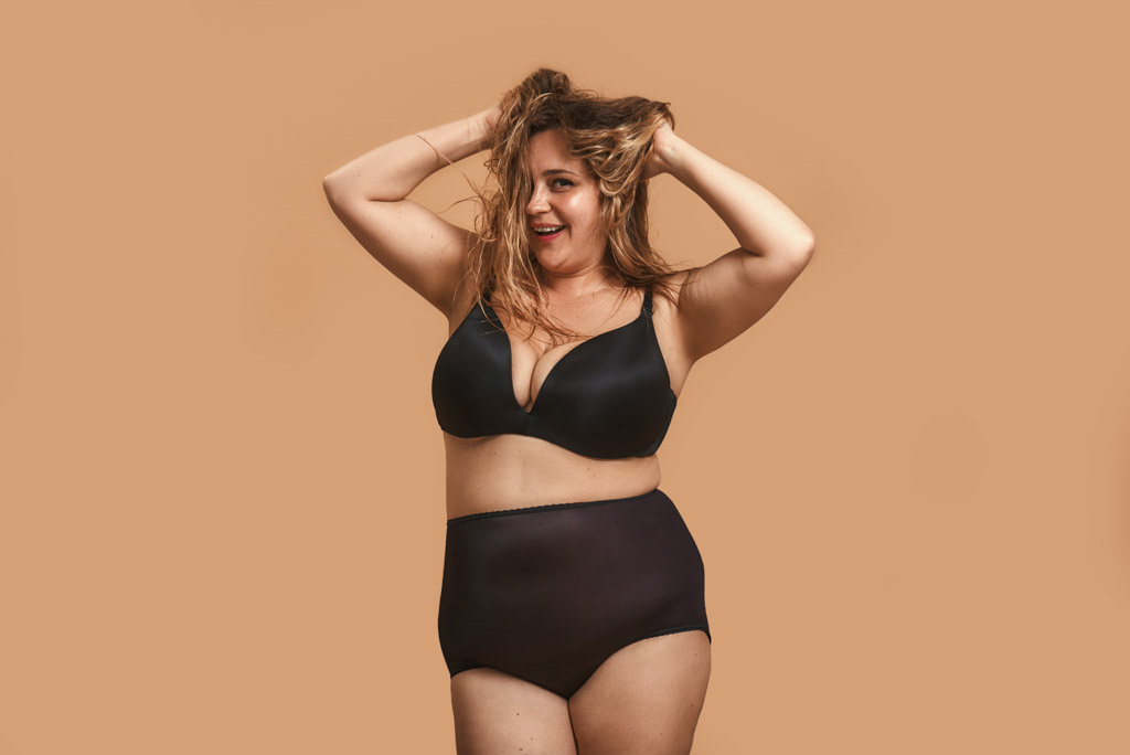Simply Happy. Positive Plump Woman In Black Underwear Touching Her Hair And Laughing While Standing Against Brown Wall In Studio
